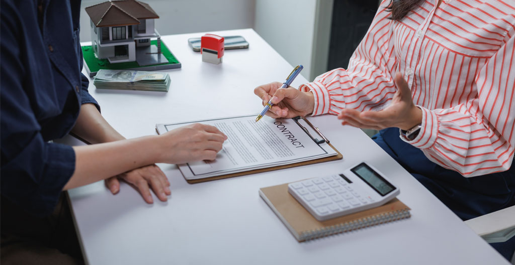 Gros plan sur des mains signant des documents officiels sur un bureau en bois clair avec un stylo et un tampon de notaire visible au premier plan